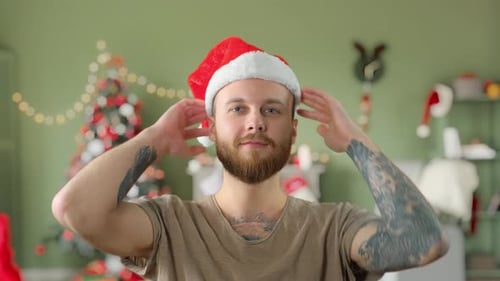 Young Handsome Bearded Tattooed Guy in Green T-shirt Is Put on a Red Christmas Hat and Smiling at