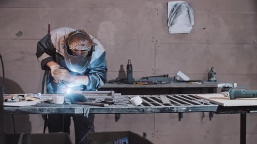 Junger Mann Arbeiter in Schutzhandschuhen und Helm, der Teile des Metalldetails zusammenschweißt