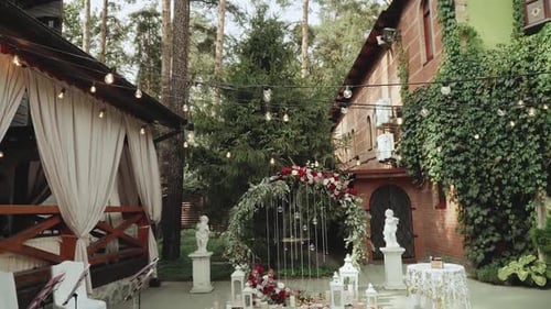 Wedding Arch with Red Flowers and White Chairs Lamp Garland Over the Ceremony Near Restaurant Slow