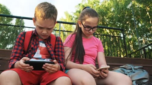The Older Sister and Brother Are Sitting on the Street, Looking at Smartphones.