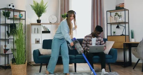 Chica con auriculares limpiando alfombra con aspiradora mientras su novio de piel negra