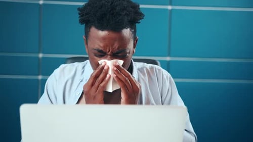 Sick Young African American Man Sneezes Covering Nose with Paper Tissue