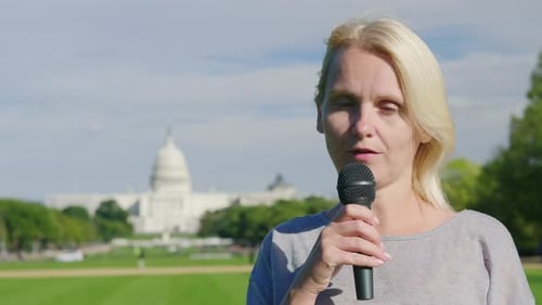 A Young Female Reporter Tells the News in a Microphone Against the Backdrop of the Capitol Building