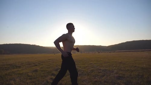 Young Muscular Man Running Through Field with Beautiful Landscape at Background. Male Athlete Trains