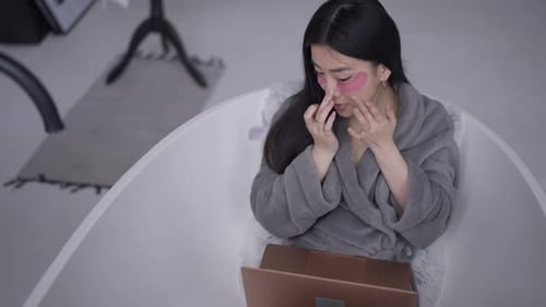 High Angle View of Young Slim Asian Woman Applying Eye Patches Looking at Laptop Screen Sitting in
