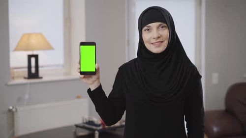 Portrait of Smiling Muslim Woman in Hijab Holding Smartphone with Greenscreen. Modern Eastern Lady