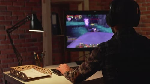 Rückansicht eines kaukasischen Mannes, der im Kopfhörer sitzt und im Licht einer Tischlampe ein Computerspiel spielt