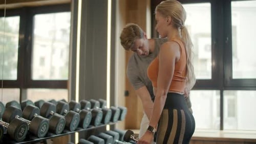 Sporty Fit Girl Making Bicep Curls in Club. Trainer Correcting Client at Gym