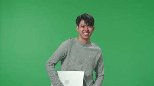 A Smiling Asian Man Holding Computer While Standing In Front Of Green Screen Background