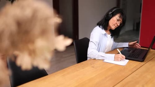 Woman at Table with Laptop Writing in Notebook
