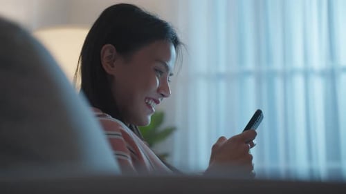 Asiatische schöne Frau sitzt auf dem Sofa und unterhält sich nachts mit dem Handy.
