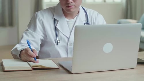 Male Doctor Working With Laptop And Writing On Notebook At Home Office