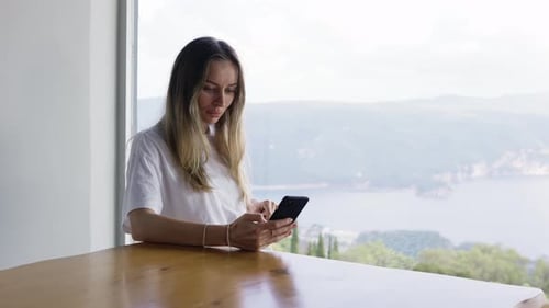 Girl Surfing Internet at Her Phone While Sitting with the Panoramic Window Landscape View