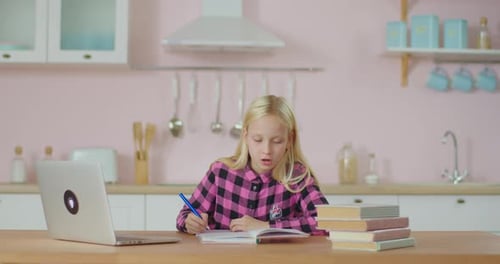 School Kid Writing in Notebook While Studying Online on Laptop at Home. Blonde 10 Years Old Girl