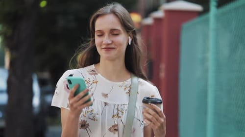 Smiling Young Woman Walks with Phone and Coffee