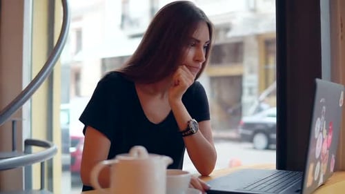 Young Woman Sitting in the Coffee Shop By the Window Having a Coffee Break She is Using Her Laptop