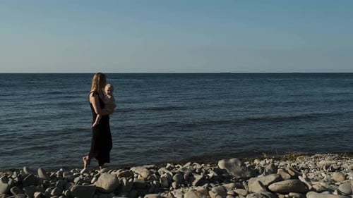Mom in a Black Dress with a Child in Her Arms Goes Forward Along the Beach. Endless Ocean on the