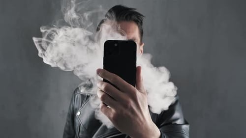 Raucher Mann in Lederjacke Versteckt sein Gesicht mit Smartphone und bläst Rauch über Dunkelgrau
