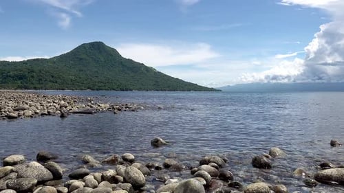 felsenbedeckte tropische Inselküste mit sanft vorbeiziehenden Wellen und hohem, baumbedecktem Berg in