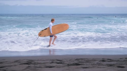 Surfer Walks Along Dark Sand Beach with Surfboard
