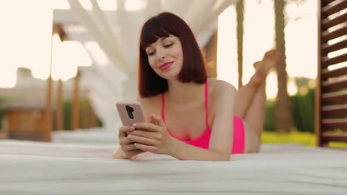 Focused Young Caucasian Lady in Pink Swimsuit Lying in a Wooden Gazebo Bed Near Resort Pool