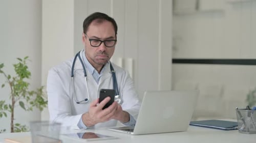 Middle Aged Doctor Using Smartphone While Working on Laptop in Office