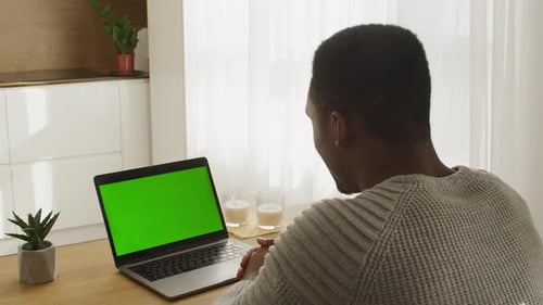 Young African American Man Sitting in Front of a Green Screen Laptop on a Video Call in a Sunny