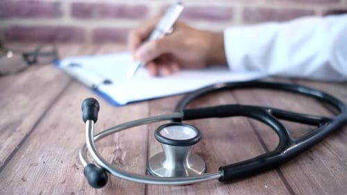 Stethoscope and Doctor Hand Writing Prescription on Clinic Table .