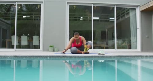 Happy african american plus size woman practicing yoga, using smartphone next to swimming pool