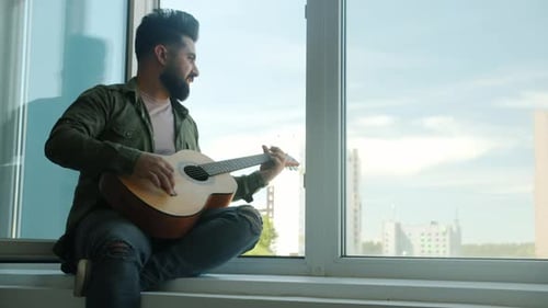 Man Playing Guitar by Apartment Window in Daytime