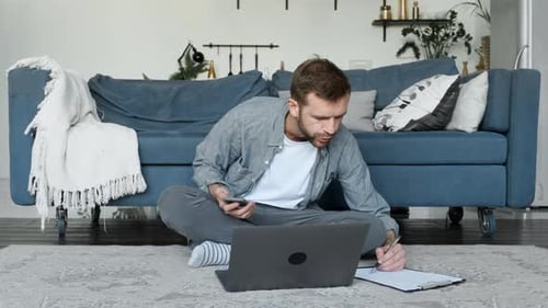 A Young Man Takes Notes on Paper, A Freelancer Works At Home, Uses A Laptop Computer