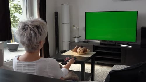 A Middle-aged Woman Sits, Watches TV with a Green Screen and Switches Programs with a Remote Control
