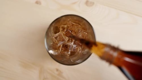Top View of Cola Sparkling Drink Pouring in a Glass with Ice Cubes on Light Brown Wooden Table