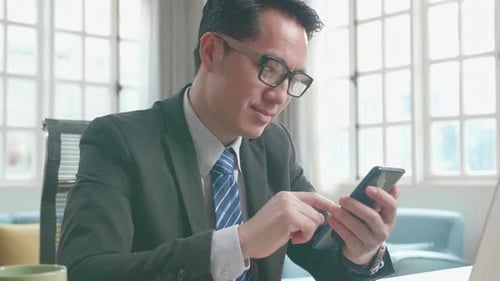 Asiatischer Geschäftsmann benutzt Handy, während er den Computer für die Arbeit zu Hause benutzt