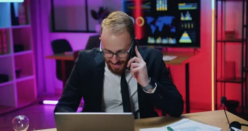Businessman Talking on Phone with Colleague or Client and Looking at Laptop Screen in Evening office
