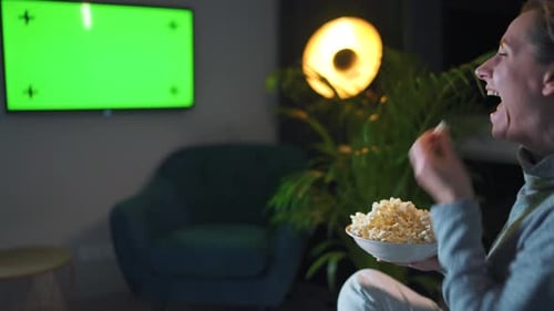 Rear View of a Woman Sitting on a Sofa in the Living Room in the Evening and Watching a Green TV