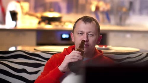 In the evening, sitting at the TV on the sofa, a young man drinks beer, camera movement