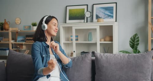 Câmera lenta de menina em fones de ouvido dançando ouvindo música sentada no sofá