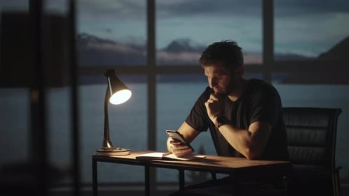 Silhouette of a Man Sitting at a Table Looking at a Mobile Phone Thinking and Analyzing the Stock