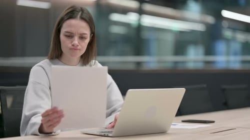 Woman Reviews Document While Using Laptop in Office