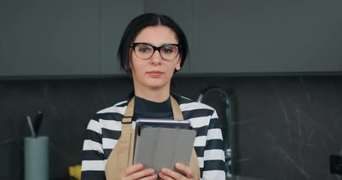 Retrato de mujer concentrada ocupada atractiva en delantal de cocina sosteniendo dispositivo tableta