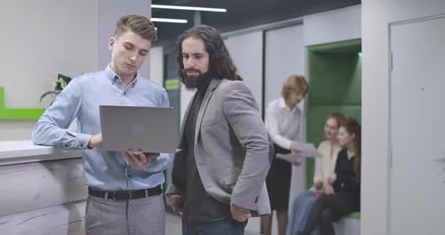 Young Caucasian and Middle Eastern Men Standing with Laptop in Open Space Office and Talking