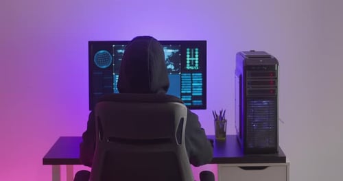 Rear View of Anonymous Hacker in Hood Working at Computer in a Data Center at Night