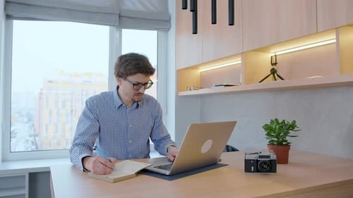 Man in glasses works at a laptop against the background of a window. Freelancer working remotely.