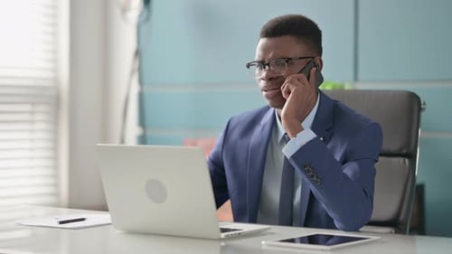 Angry Young African Businessman Talking on Phone While Using Laptop in Office