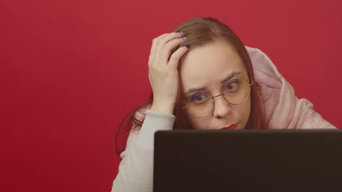 Young Woman in Glasses Works on Laptop and Scratches Head Sitting in Home Office