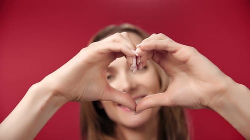 Closeup Smiling Young Woman Making Sign Heart Shape By Hand Posing at Red Studio Background