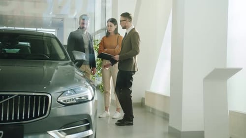 Happy Young Family Husband and Wife Choosing Auto in Car Dealership Talking To Salesman