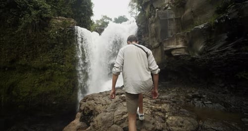 Tracking Handheld Close Up Shot of a Young Man Walking Up the Rocky Floor Towards a Large Waterfall