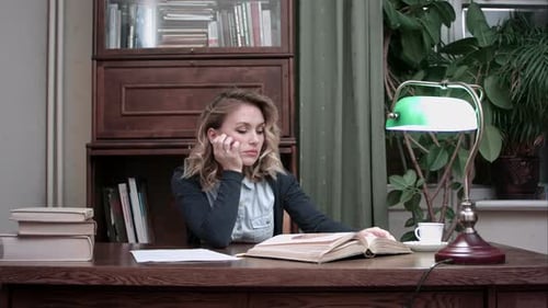 Tired Young Woman Falling Asleep Over a Book While Sitting at the Table